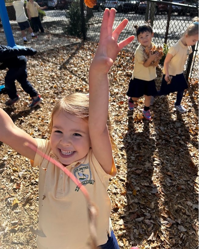 Pre-K student stands on the playground, cover with fall leaves, her hands in the air and a smile on her face