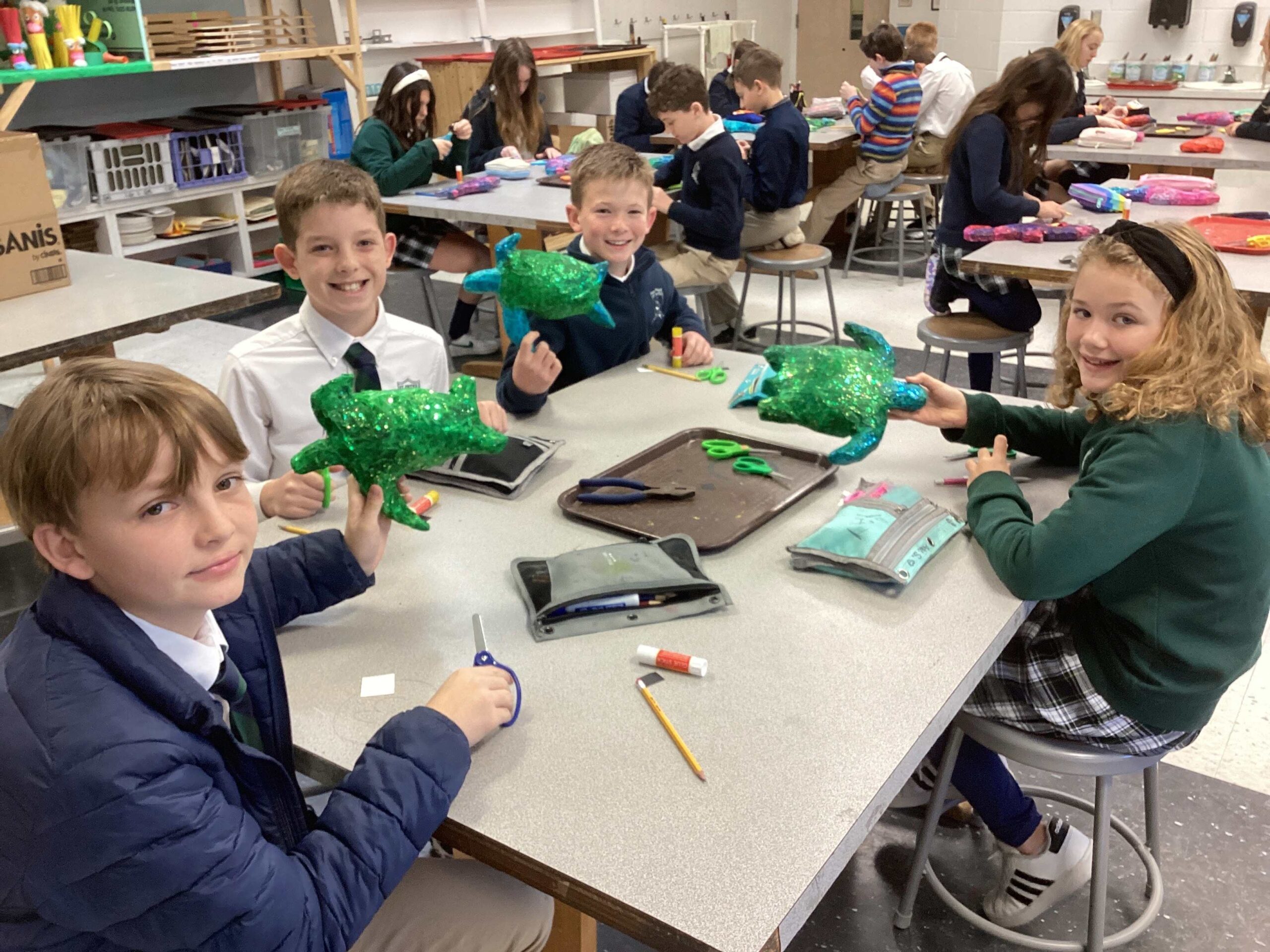 A group of students sit at the art table, smiling as they show their large, green glittered 3D turtles
