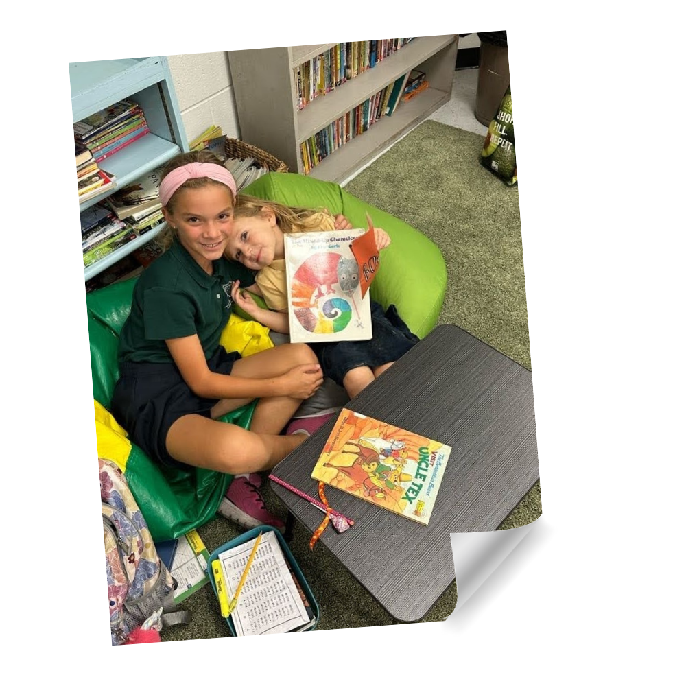 Student mentor book buddy sitting with younger student on bean bag chairs in the library, reading a book together