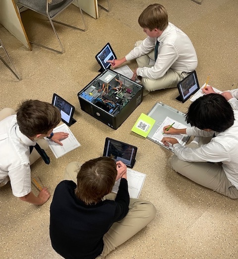 Students sit on the floor with an open computer hard drive in front of them, working on worksheets and iPads