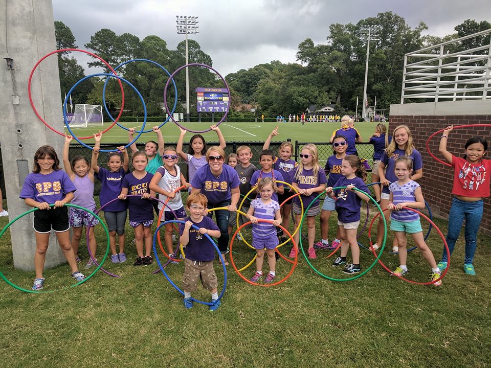 Large group of elementary student members of the Hoopsters and their adult leader, stand outside the playing field gate, holding up their hula hoops in the air