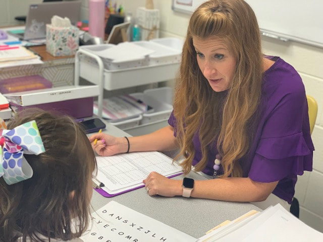 Woman works with child at a desk filling out an evaluation while the student reads letters from a paper