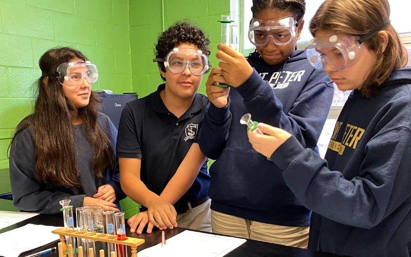 Middle school students stand looking at a beaker with clear goggles on, in science class